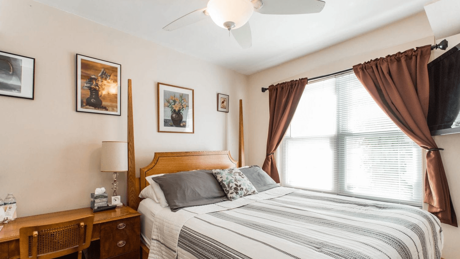 A cozy bedroom featuring a wooden bed, decorative pillows, artwork on the walls, and a window with brown curtains.