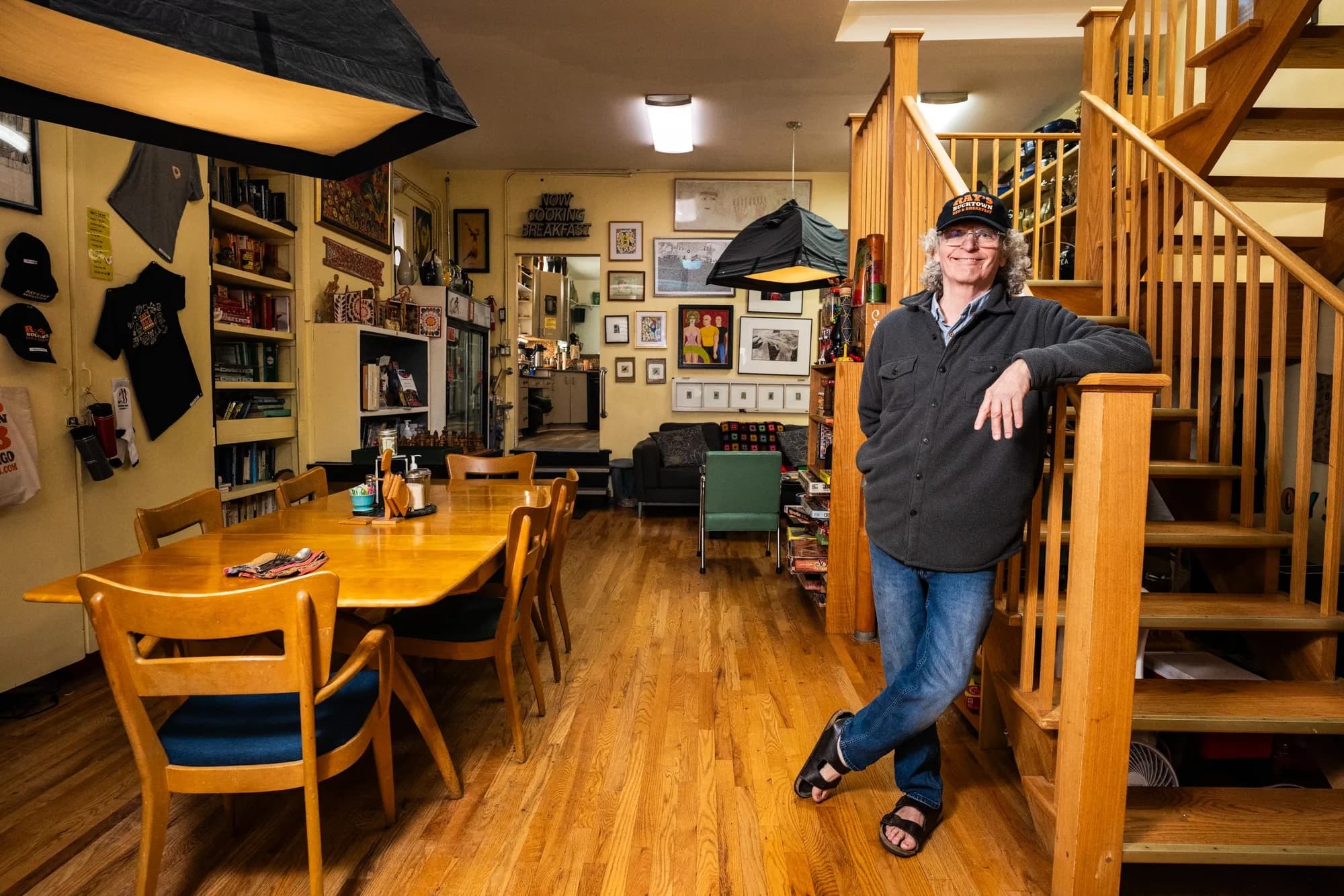 A person stands casually by a wooden staircase in a cozy, art-filled dining area.