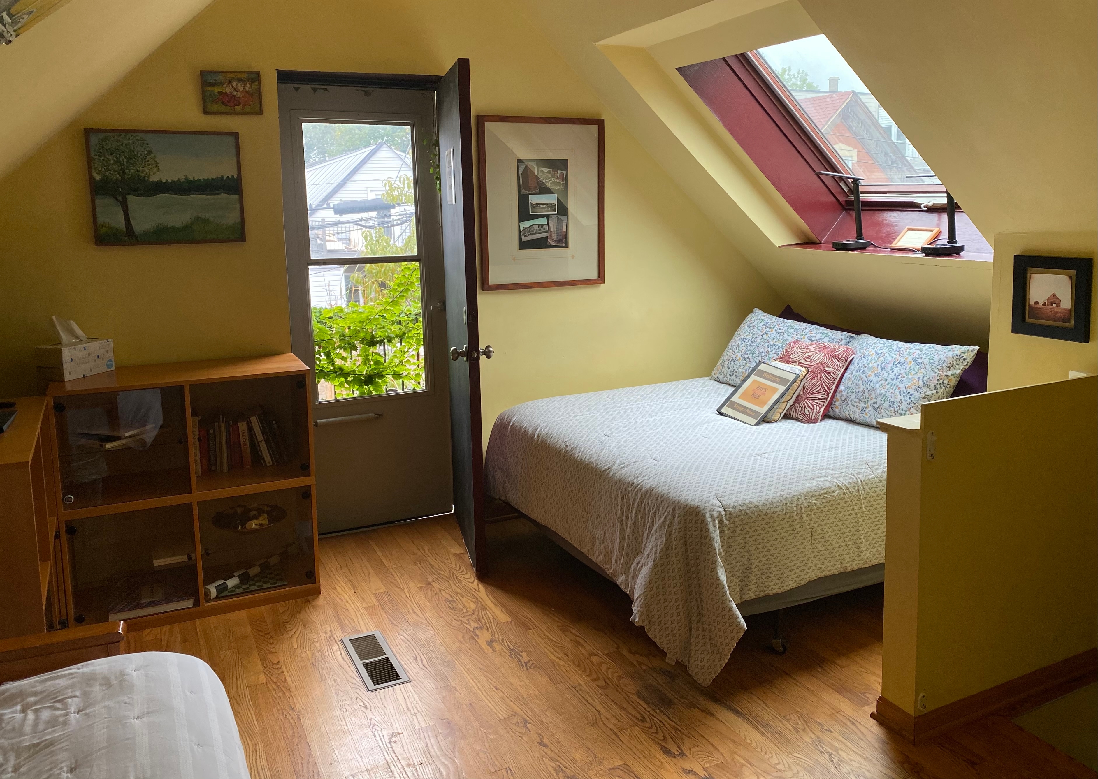 room with angled a frame ceiling painted tan with queen bed with brown comforter, and futon couch with dark brown cover. Blond wood shelves, oak floors, door that goes out to roof top deck and skylight.