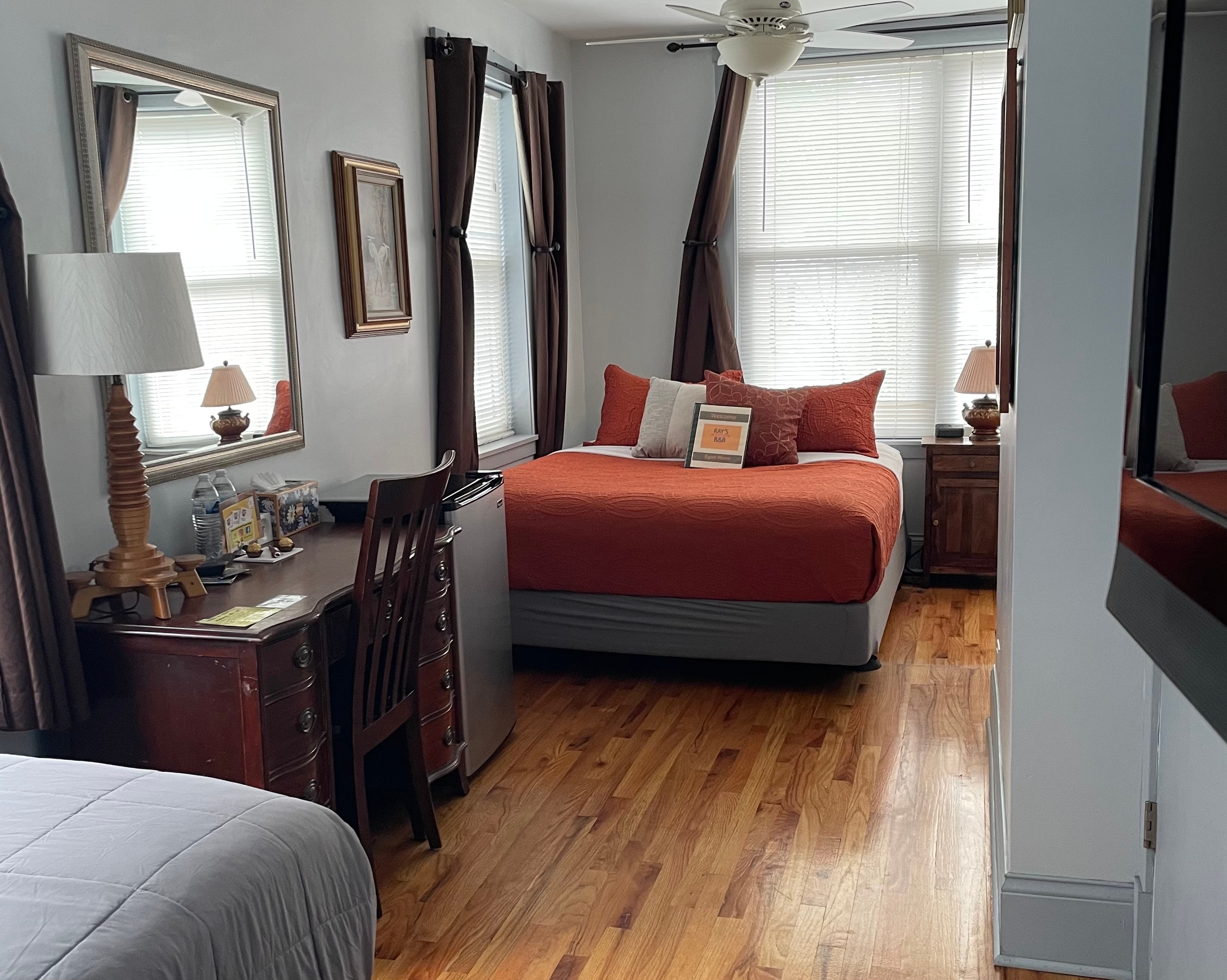 queen bed with grey pattern comforter, desk and 4 large double hung windows with blinds