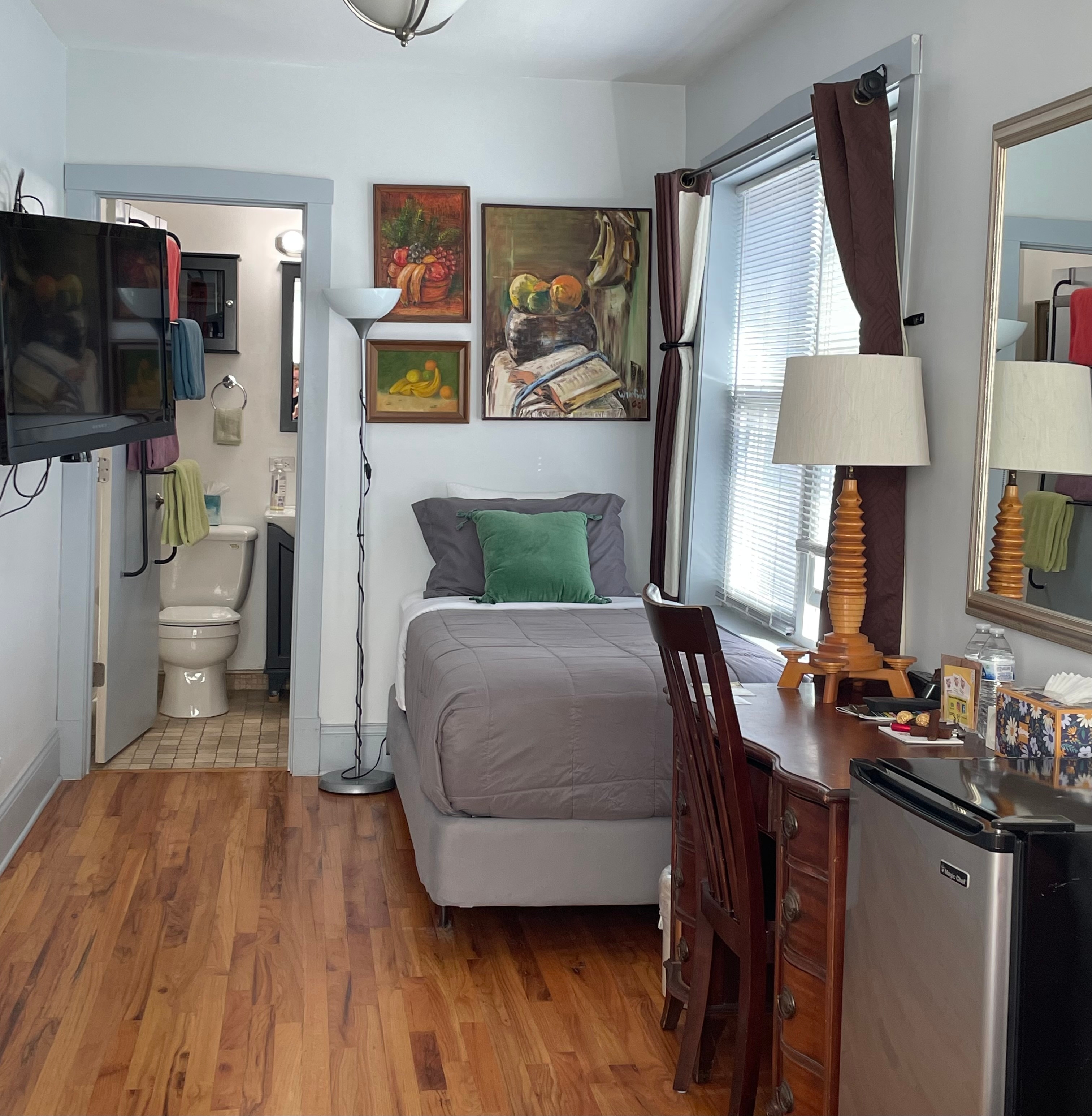 twin bed with tan & brown comforter, HDTV on wall, two double hung white windows with blinds, and door open into en-suite private bathroom