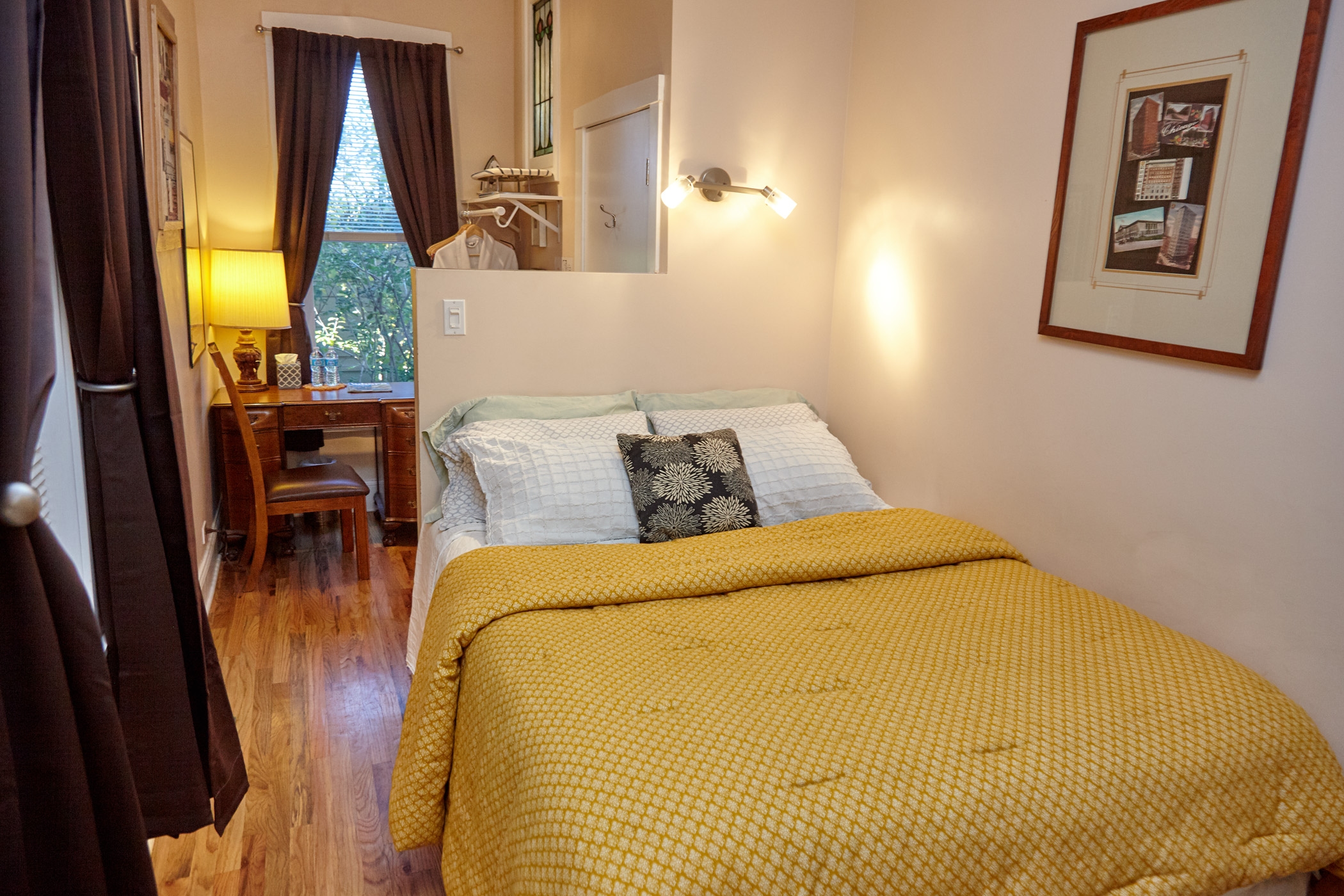 queen bed with tan comforter, tan walls, 4 pillows, wooden desk and two double hung windows.