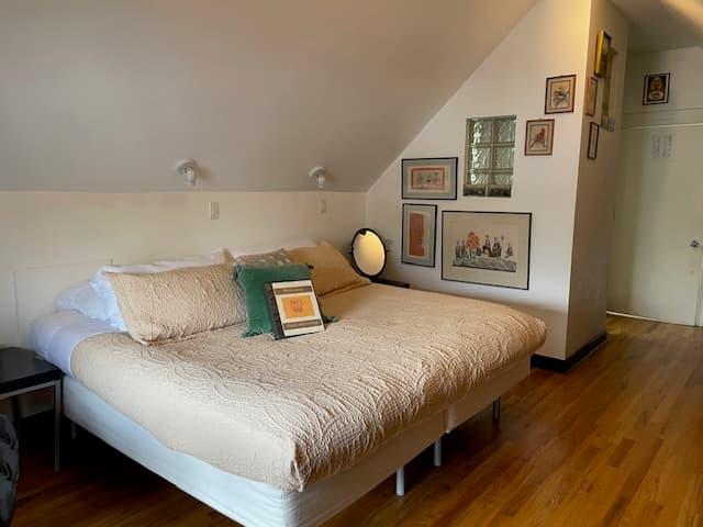 king bed with green comforter, two padded rolling chairs, oak floors, 2 double-hung and one picture windows.